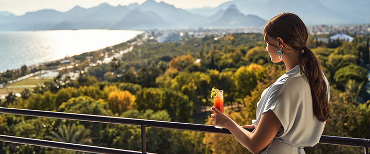 Eine Frau lehnt am Geländer, hält einen Cocktail in der Hand und schaut über eine grüne Palmenlandschaft mit Meerblick und Bergkulisse.